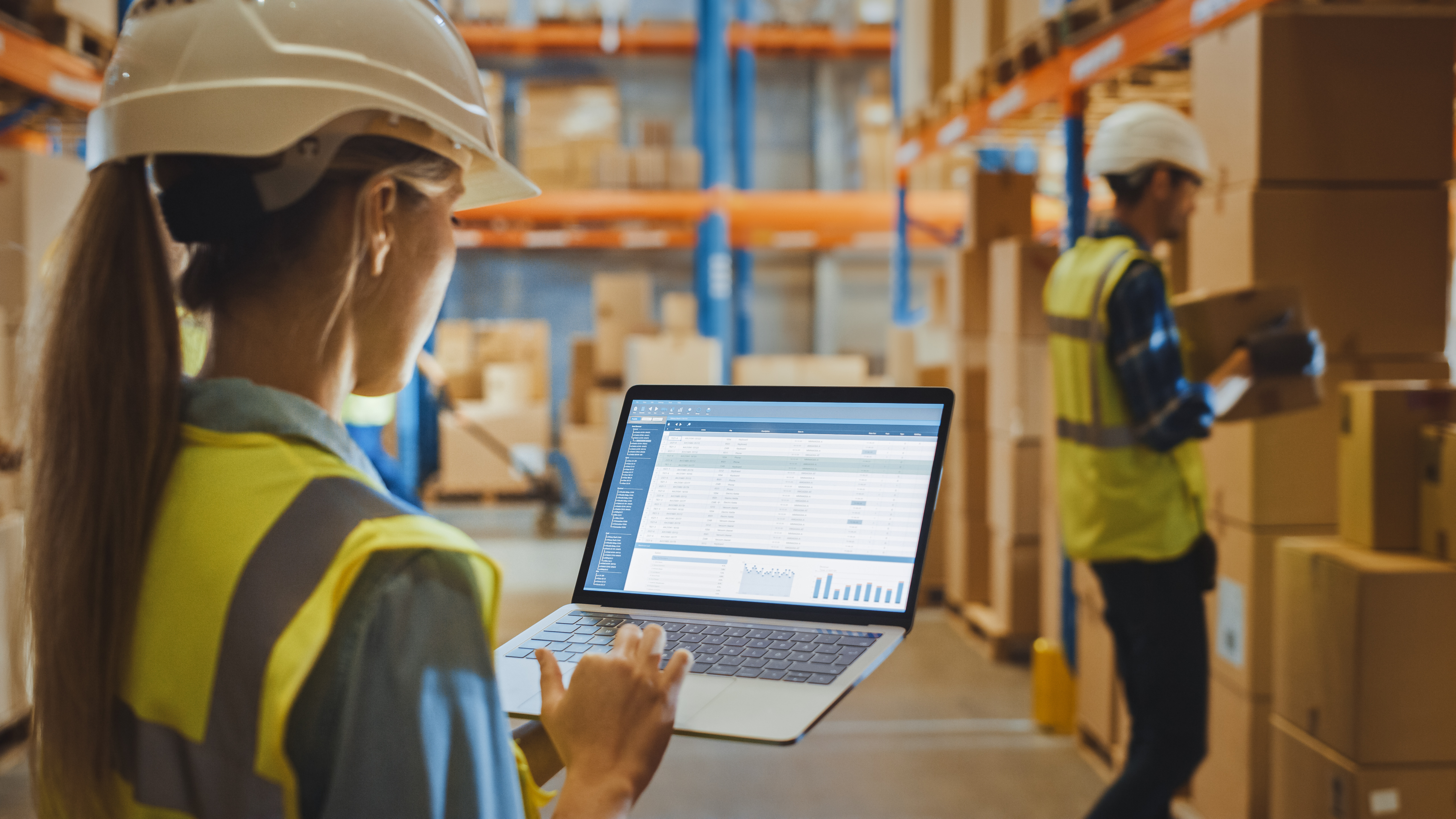 Een vrouwelijke magazijnmedewerker met een veiligheidshelm houdt een laptop vast en bekijkt een rapport over Warehouse Profitability.